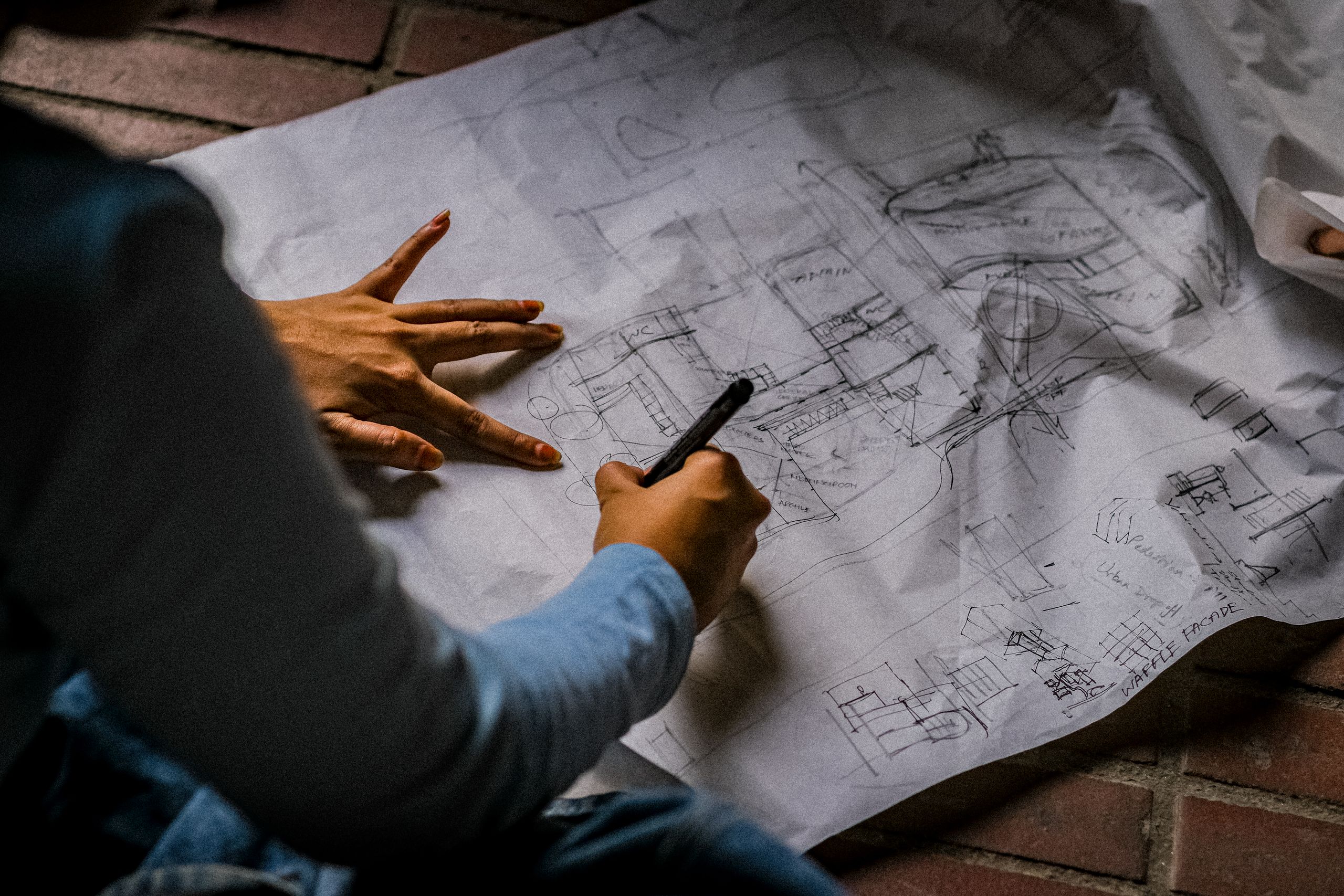 Person sketching architectural design on blueprint paper with pen on brick floor.
